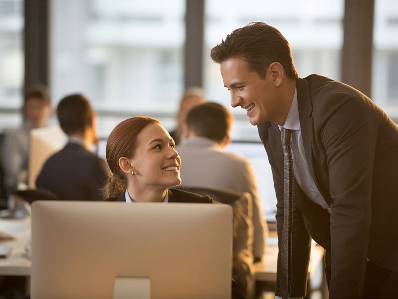 A professional man leans over a desk smiling at a woman seated at her computer in a bright office, suggesting focused personal attention.