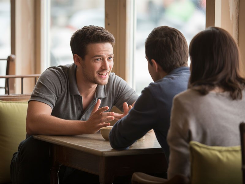 A man sitting at a café table talking animatedly with two friends, appearing cheerful and engaged in conversation.