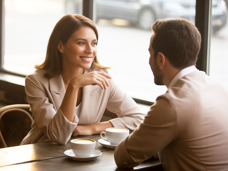 A smiling woman and man enjoying a friendly conversation over coffee at a cozy café table near a large window.