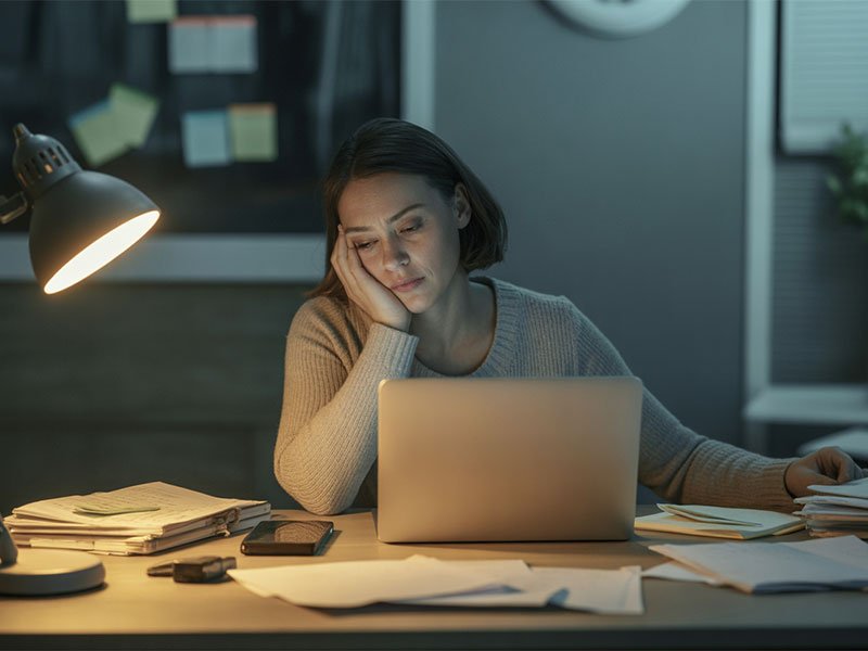 A tired woman sitting at a desk late at night, resting her face on her hand while working on a laptop under a desk lamp.
