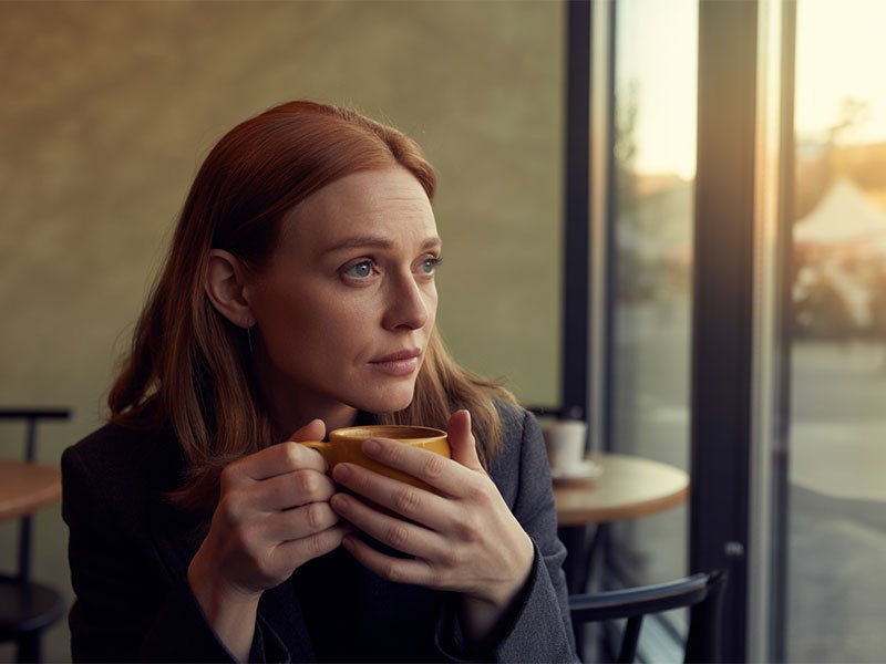 A woman sitting in a café near a window, holding a cup of coffee with both hands and looking pensively outside.