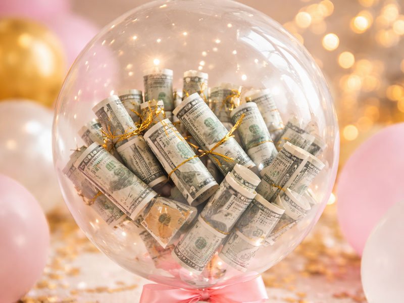 Clear balloon filled with rolled dollar bills tied with gold string, displayed as a creative cash gift with a festive background.