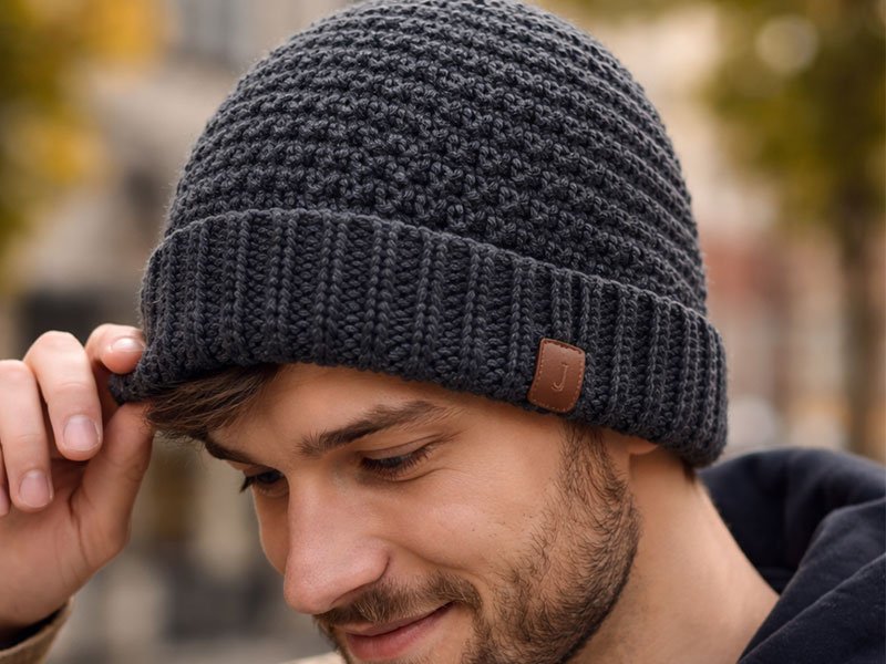 Man wearing a dark gray crochet beanie, smiling slightly outdoors in cool weather.