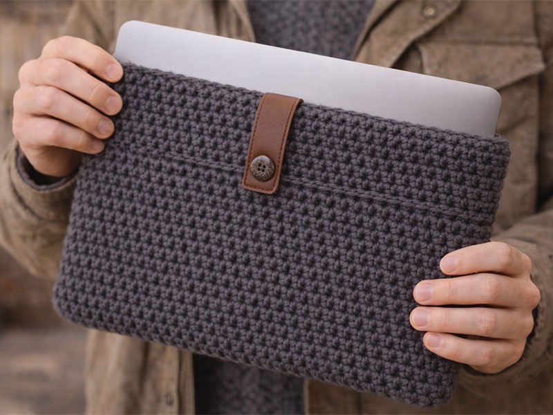 Person holding a gray crochet laptop sleeve with a button strap, protecting a laptop in a handmade case.