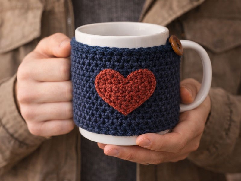 Person holding a coffee mug wrapped in a blue crochet mug cozy with a small heart design.