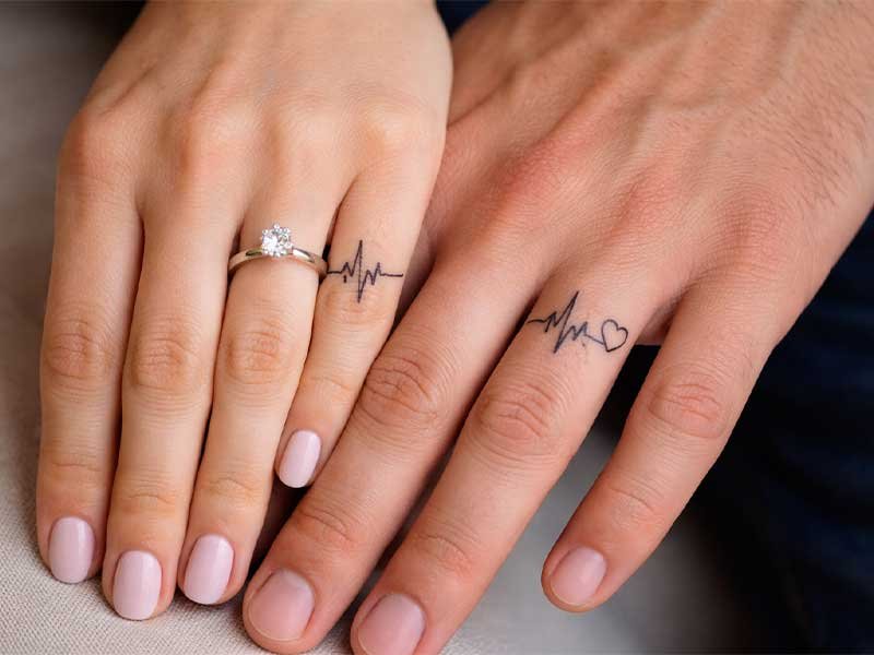 Close-up of two hands showing matching heartbeat and heart tattoos near the ring fingers, with wedding ring.