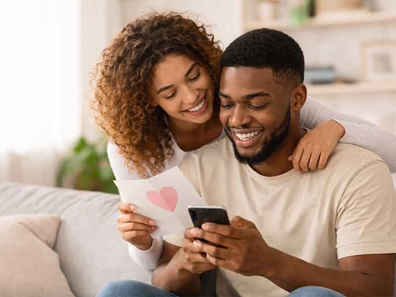 A couple sitting together, with the woman holding a love note and the man smiling as he looks at his phone.