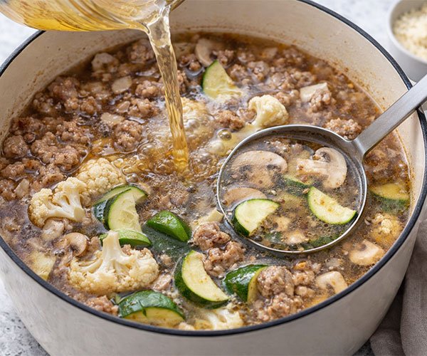 Chicken broth being poured into a pot with Italian sausage and low-carb vegetables for keto sausage soup.