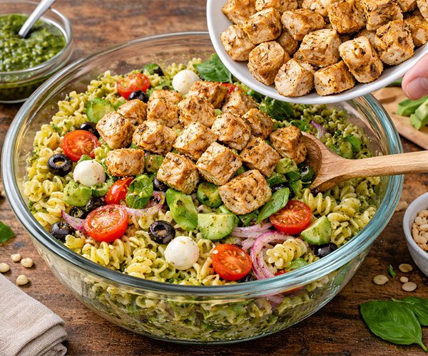 Cooked chicken being added and mixed into a large bowl of pesto pasta salad with vegetables and cheese.