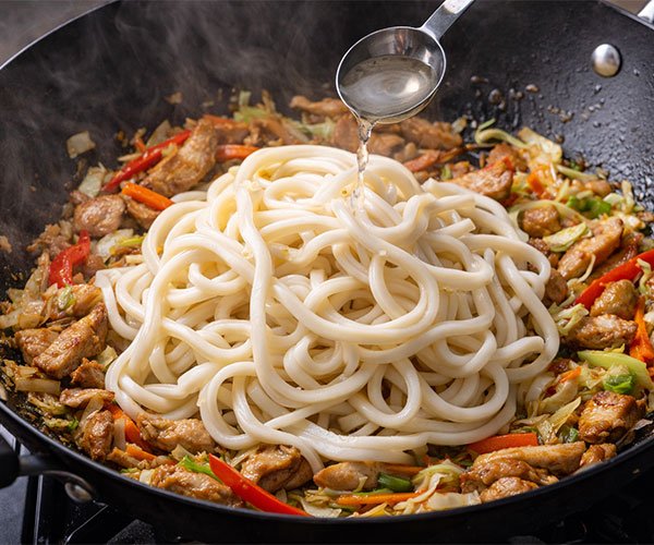Udon noodles added to a hot wok with stir-fried chicken and vegetables, with water being poured in to loosen the noodles.