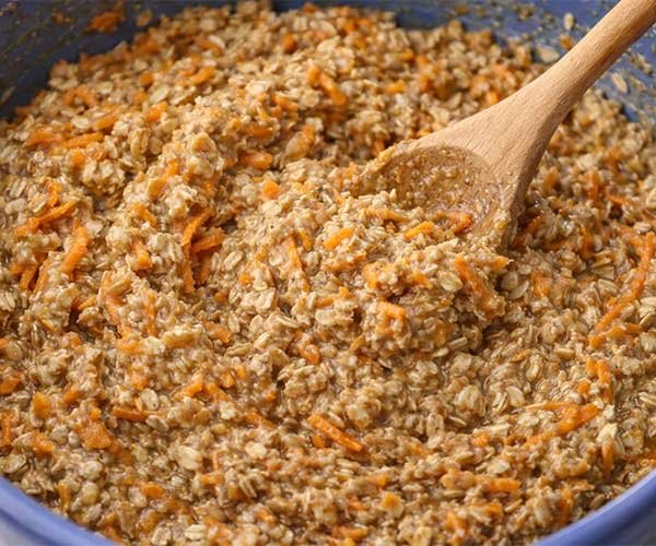 Carrot cake oatmeal batter with grated carrots and oats being mixed in a bowl with a wooden spoon.