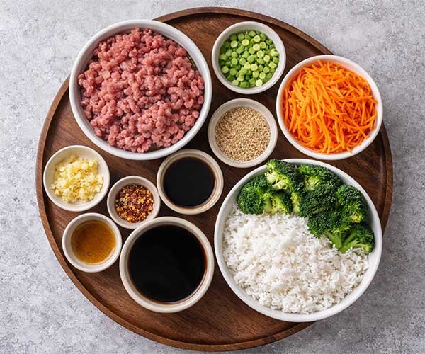Ingredients for Korean ground beef bowl on a brown tray, including ground beef, rice, soy sauce, sesame oil, garlic, sesame seeds, green onions, carrots, and broccoli.