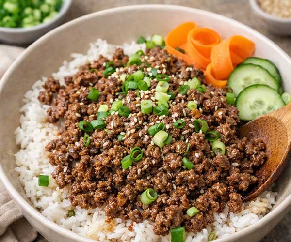 Korean ground beef bowl served over white rice, topped with green onions, sesame seeds, cucumber slices, and carrot ribbons.