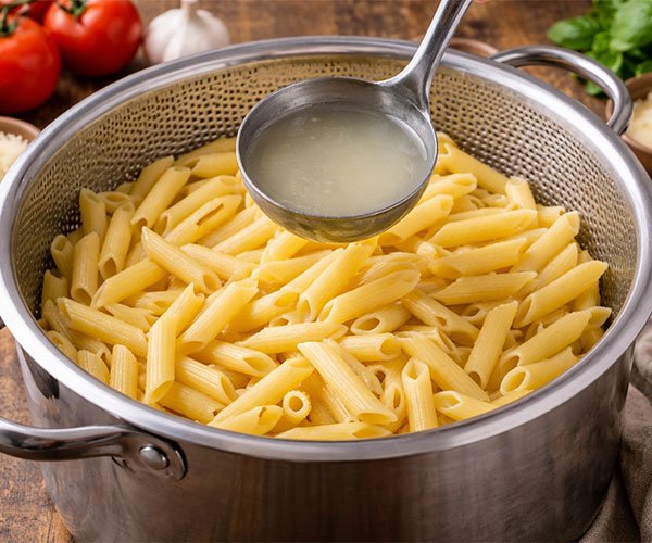 Cooked penne pasta in a colander with a ladle of reserved pasta water, with tomatoes, garlic, basil, and parmesan in the background.