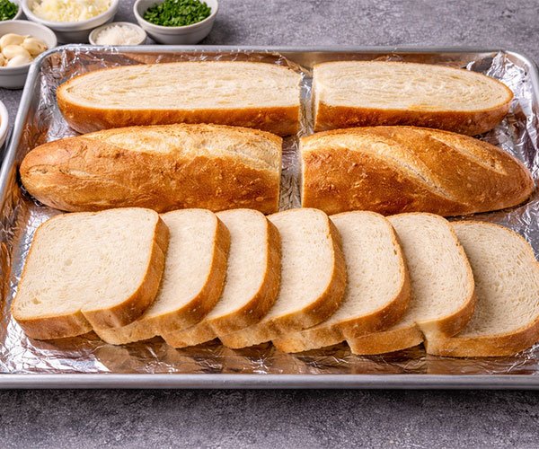 Sliced bread arranged on a foil-lined baking sheet, ready for cheesy garlic bread toppings, with bowls of cheese, garlic, and herbs nearby.