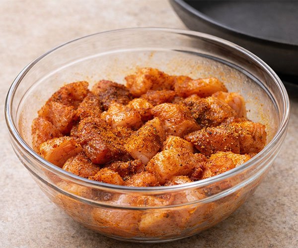 Seasoned chicken pieces coated in spices in a glass bowl, resting on the counter with a pan heating nearby.