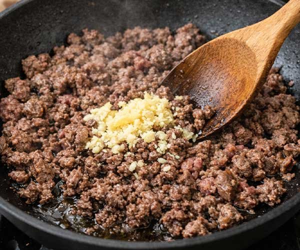 Ground beef in a skillet with minced garlic and ginger being stirred in with a wooden spoon.