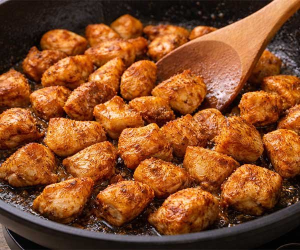 Seasoned chicken pieces sizzling in a skillet until golden, being stirred with a wooden spoon.