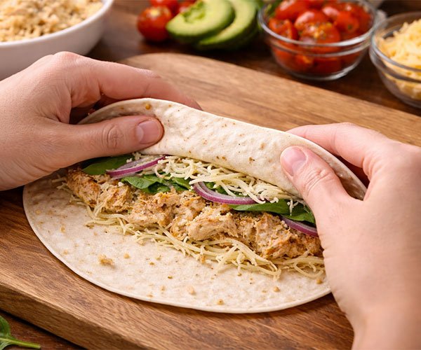 Hands folding and tightly rolling a flour tortilla filled with seasoned chicken, shredded mozzarella, fresh spinach, and red onion on a wooden cutting board, with bowls of tomatoes, avocado, and cheese in the background.