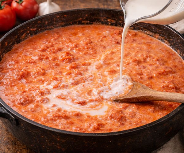 Heavy cream being poured into a simmering tomato sauce in a skillet, creating a creamy pink-orange sauce as it’s stirred.