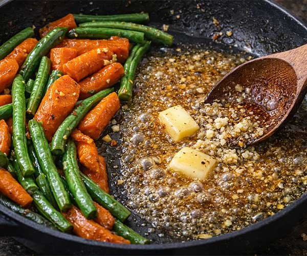 Carrots and green beans pushed to one side of a skillet while butter, honey, garlic, salt, and pepper bubble into a sticky glaze on the other side.