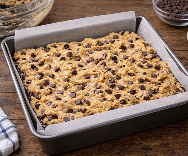 Chocolate chip cookie dough spread over brownie batter in a parchment-lined square baking pan