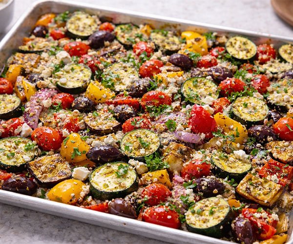 Roasted one-pan Greek vegetables with zucchini, bell peppers, cherry tomatoes, eggplant, red onion, olives, and feta, finished with fresh herbs on a sheet pan.