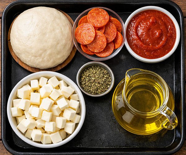 Top-down view of pizza dough, mozzarella cubes, pepperoni slices, pizza sauce, and olive oil arranged on a black tray.