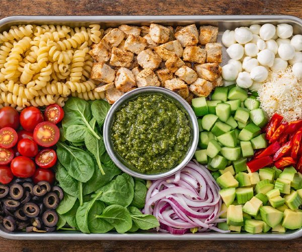 Tray filled with ingredients for chicken pesto pasta salad, including pasta, cooked chicken, pesto, vegetables, cheese, and olives.