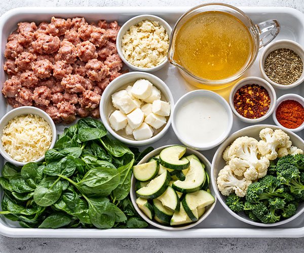 Tray of ingredients for creamy keto Italian sausage soup including sausage, broth, cream, cheese, spinach, and vegetables.