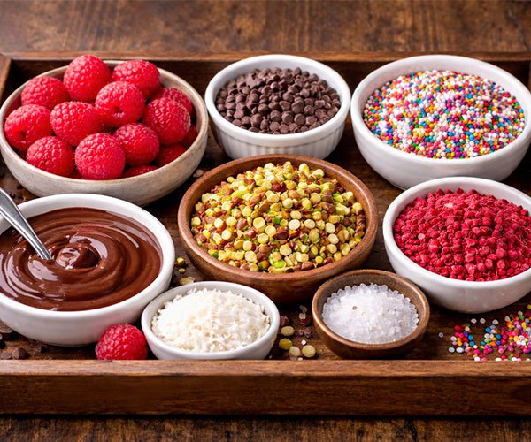 Ingredient tray with raspberries, melted chocolate, coconut flakes, chopped nuts, freeze-dried raspberries, sea salt, and colorful sprinkles.