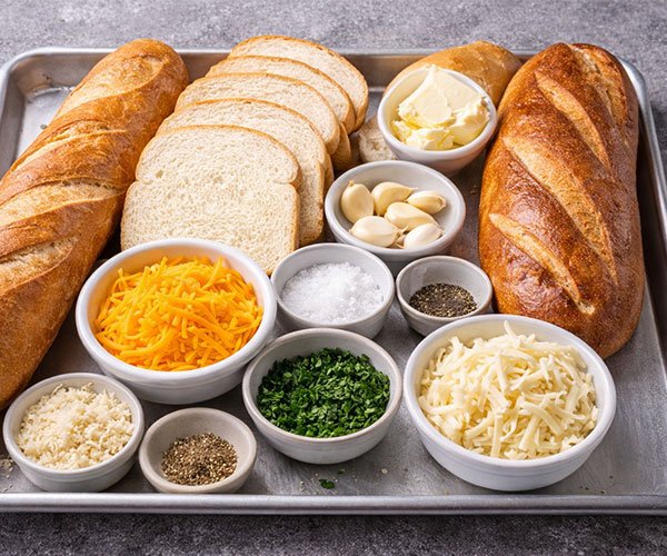 Tray of cheesy garlic bread ingredients with French bread, sliced sandwich bread, butter, garlic cloves, shredded mozzarella and cheddar, grated parmesan, parsley, salt, pepper, and Italian seasoning.