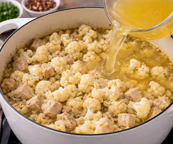 Chicken and cauliflower in a white pot as golden broth is being poured in during soup preparation