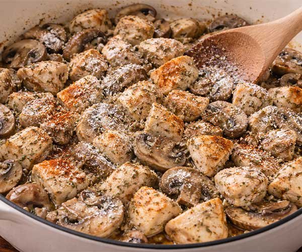 Bite-sized chicken pieces cooking with sliced mushrooms in a pot, coated in flour and seasonings like paprika, thyme, salt, and black pepper, being stirred with a wooden spoon.