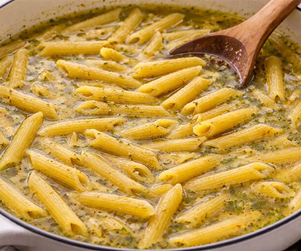 Penne pasta cooking in one pot with broth, gently simmering and being stirred with a wooden spoon.