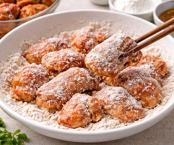 Chicken wings lightly coated in seasoned flour inside a white bowl, being lifted with chopsticks before cooking.
