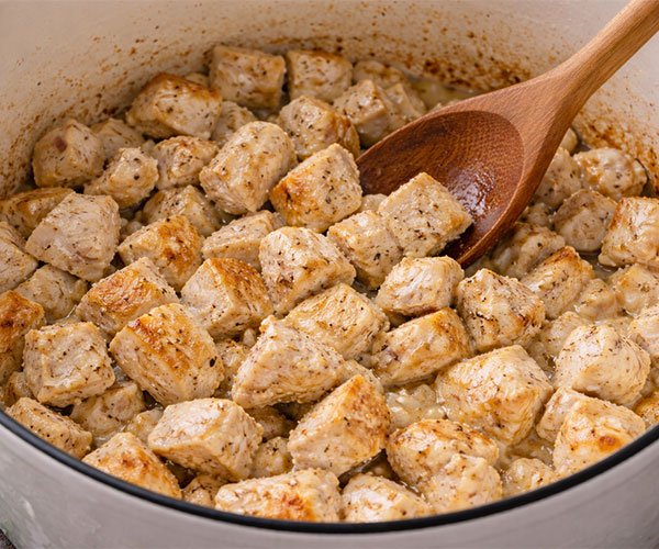 Diced chicken browning in a white pot with a wooden spoon.