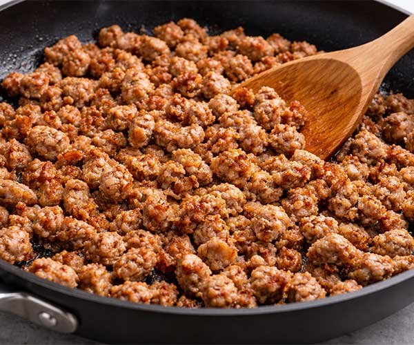 Crumbled breakfast sausage browning in a skillet while being stirred with a wooden spoon.