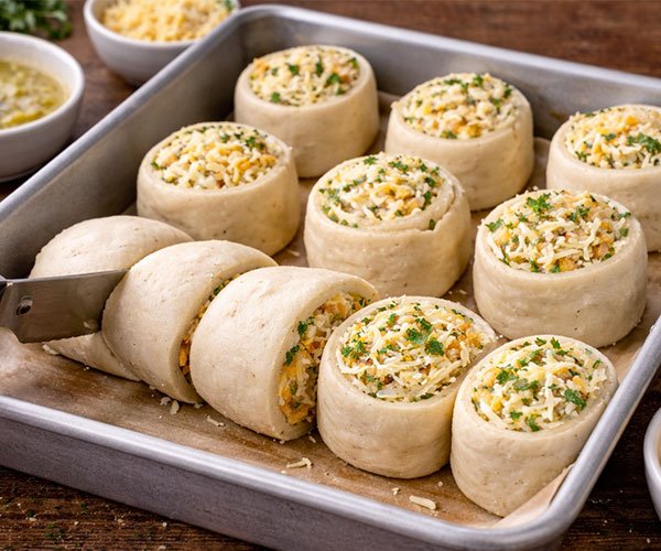Shaping garlic cheese rolls by slicing a filled dough log into even pieces and arranging them in a greased baking dish.