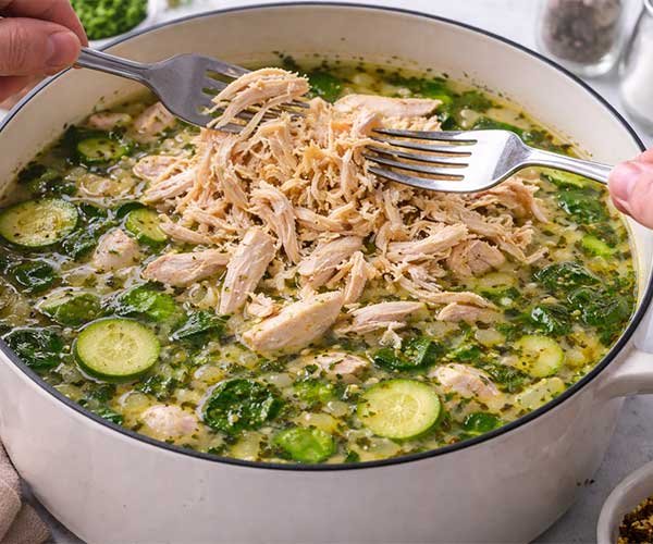 Shredded chicken being added back into a pot of keto chicken soup with vegetables and herbs.
