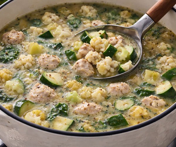 Keto chicken soup simmering with cauliflower, zucchini, and greens in a white pot with a ladle.