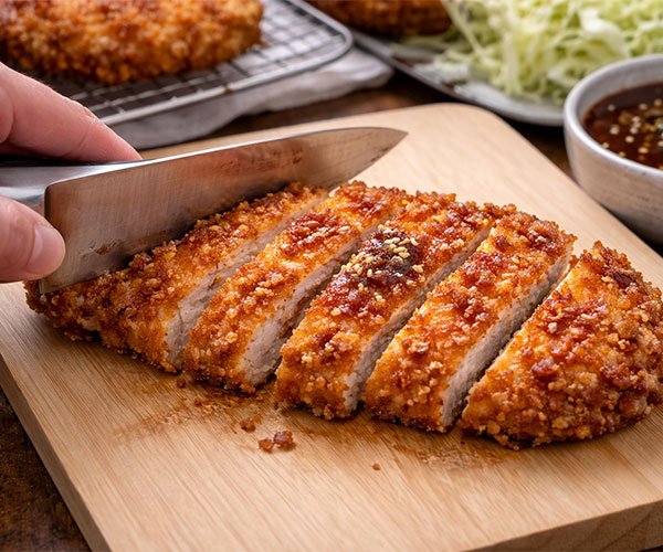 Slicing crispy Japanese katsu cutlet into thick strips on a wooden cutting board using a sharp knife.