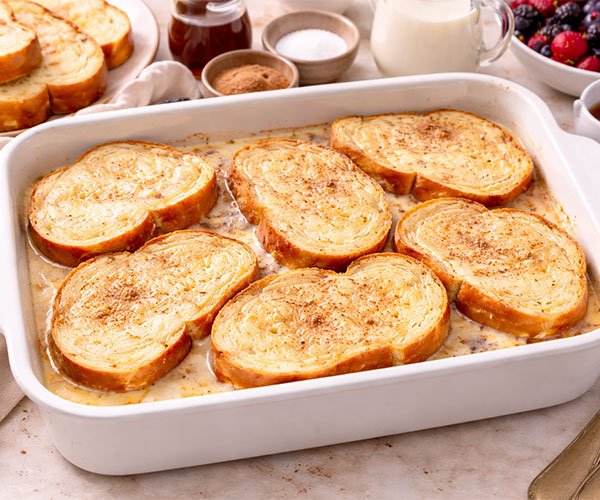 Brioche bread slices soaking in creamy custard inside a white baking dish.