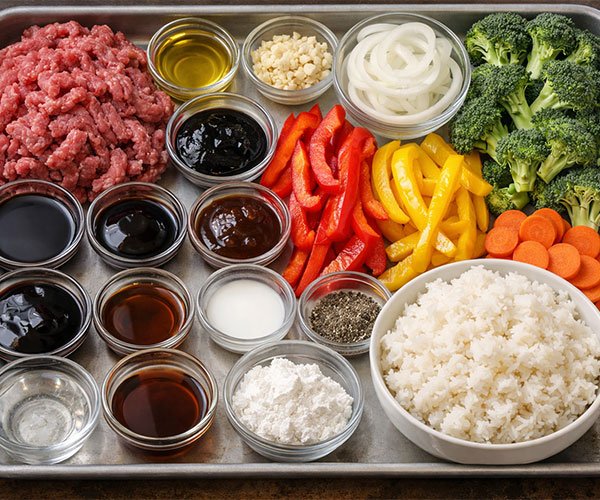 Garlic Beef Stir-Fry with Steamed Rice ingredients arranged on a metal tray, including ground beef, sliced bell peppers, broccoli, carrots, onions, garlic, sauces, cornstarch, and a bowl of steamed rice.