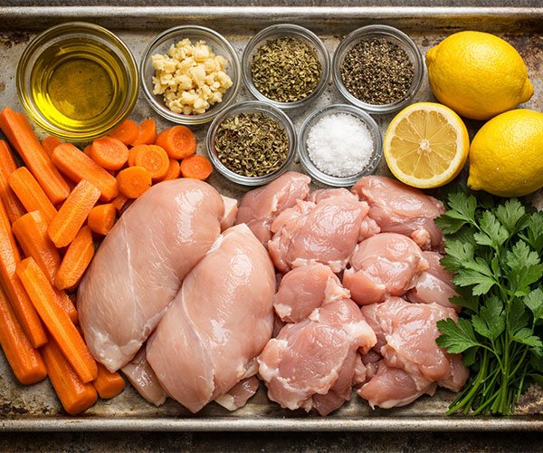 Ingredients for Juicy Garlic Herb Chicken with Roasted Carrots arranged on a tray, including raw chicken, sliced carrots, garlic, herbs, olive oil, lemon, salt, and pepper.