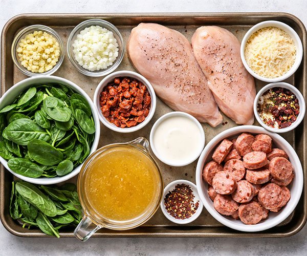 Keto Tuscan soup ingredients arranged on a tray including chicken breasts, Italian sausage, spinach, garlic, onion, Parmesan, sun-dried tomatoes, broth, cream, and seasonings.