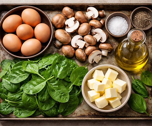 Ingredients for scrambled eggs with mushrooms and spinach arranged on a rustic tray with eggs, fresh spinach, mushrooms, butter, olive oil, salt, and pepper