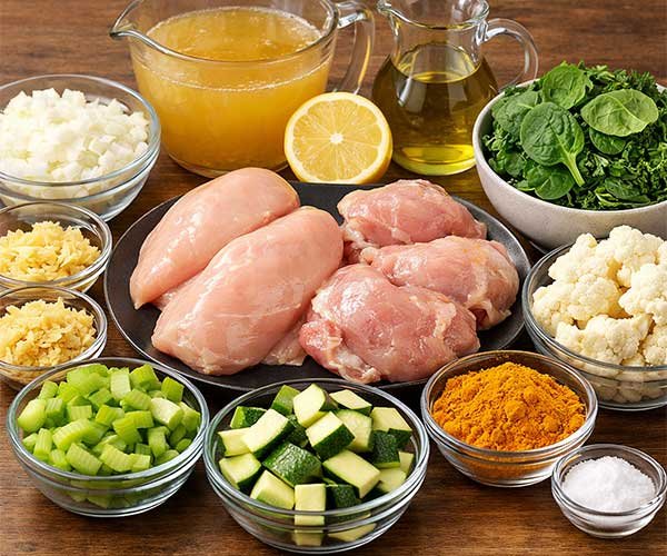 Fresh ingredients for healthy detox soup arranged in the center, including raw chicken, chopped vegetables, broth, lemon, oil, turmeric, and leafy greens.