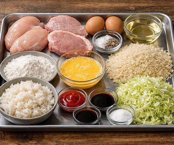 Ingredients for Japanese Katsu Bowls neatly arranged on a metal tray, including chicken breasts and pork cutlets, eggs, flour, panko breadcrumbs, rice, shredded cabbage, and sauces.