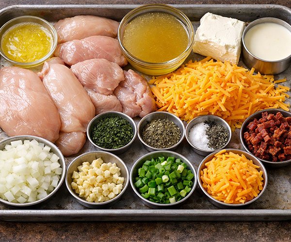 Ingredients for Keto Crack Chicken Soup arranged on a tray with raw chicken breasts, cheddar cheese, cream cheese, broth, cream, onion, garlic, herbs, and bacon.